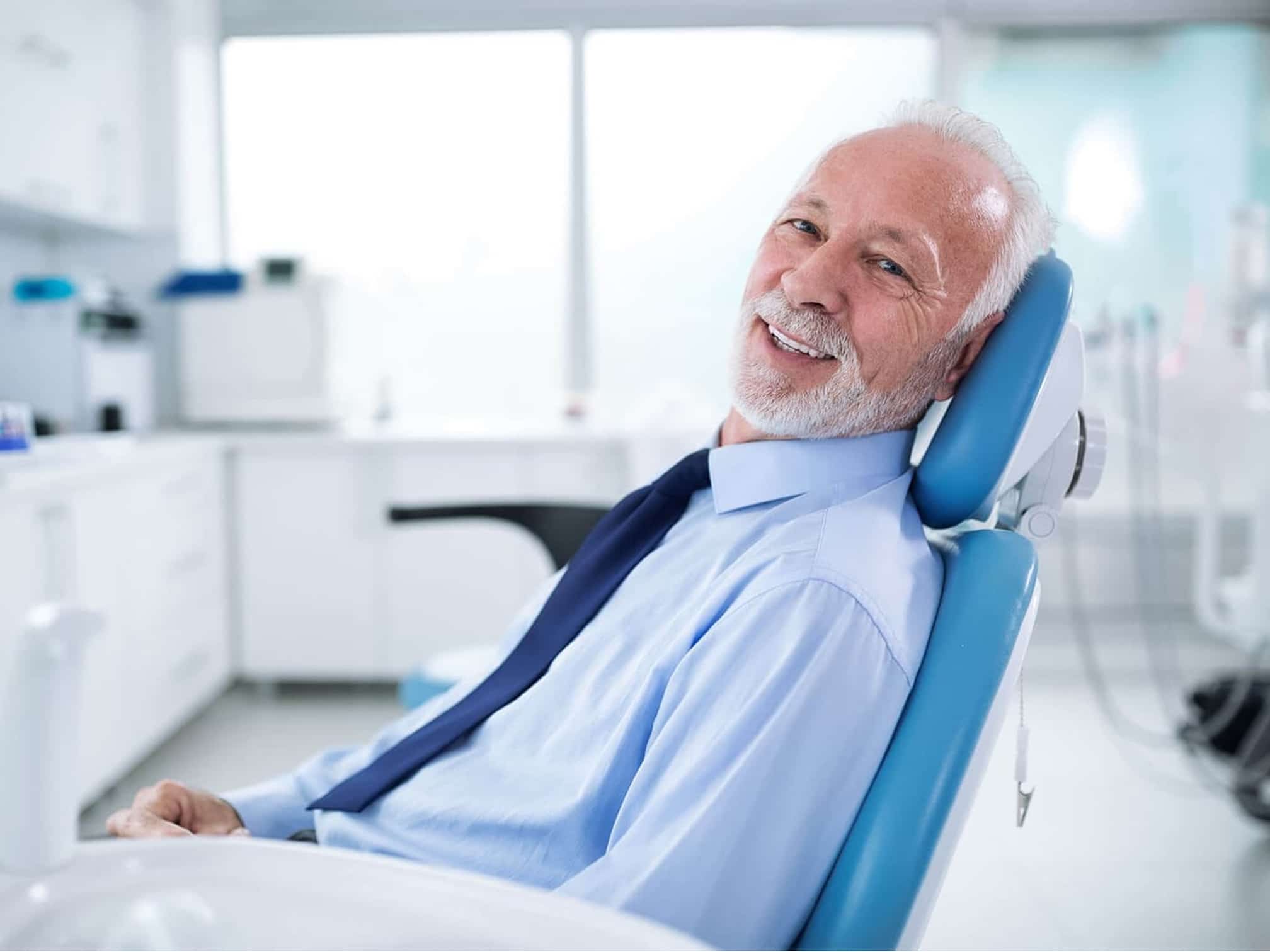 A smiling elderly man with white hair and a beard, reclines in a dental chair in a modern dental office for all on 4 dental implants in Houston